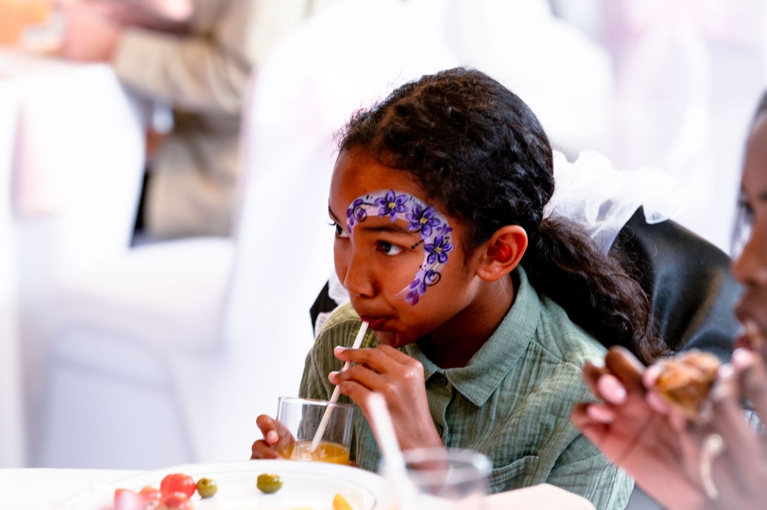 A child with face painted in purple floral designs enjoys a drink with a straw.