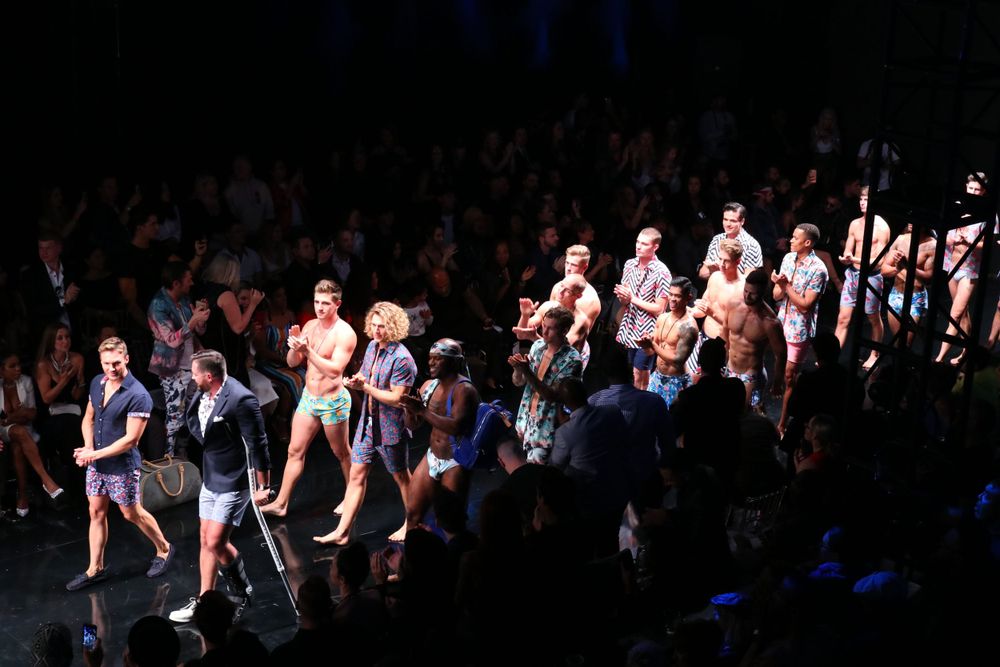 Fashion models walk the runway finale in coordinated outfits during a dark-lit fashion show.