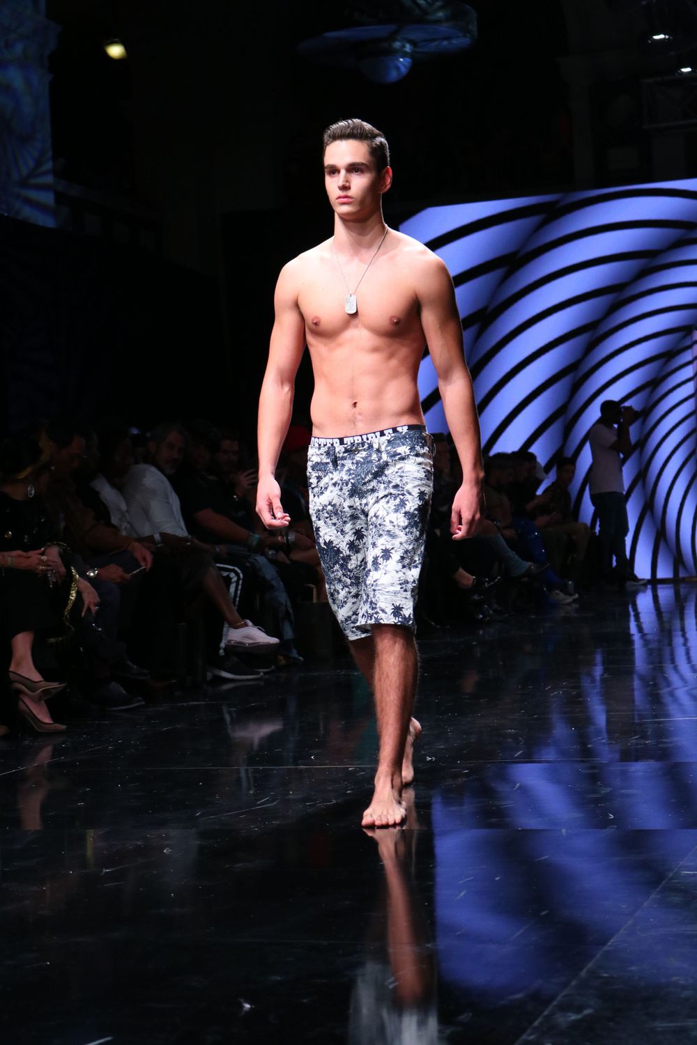 Model showcases patterned swim shorts on illuminated runway with blue lighting and artistic backdrop.
