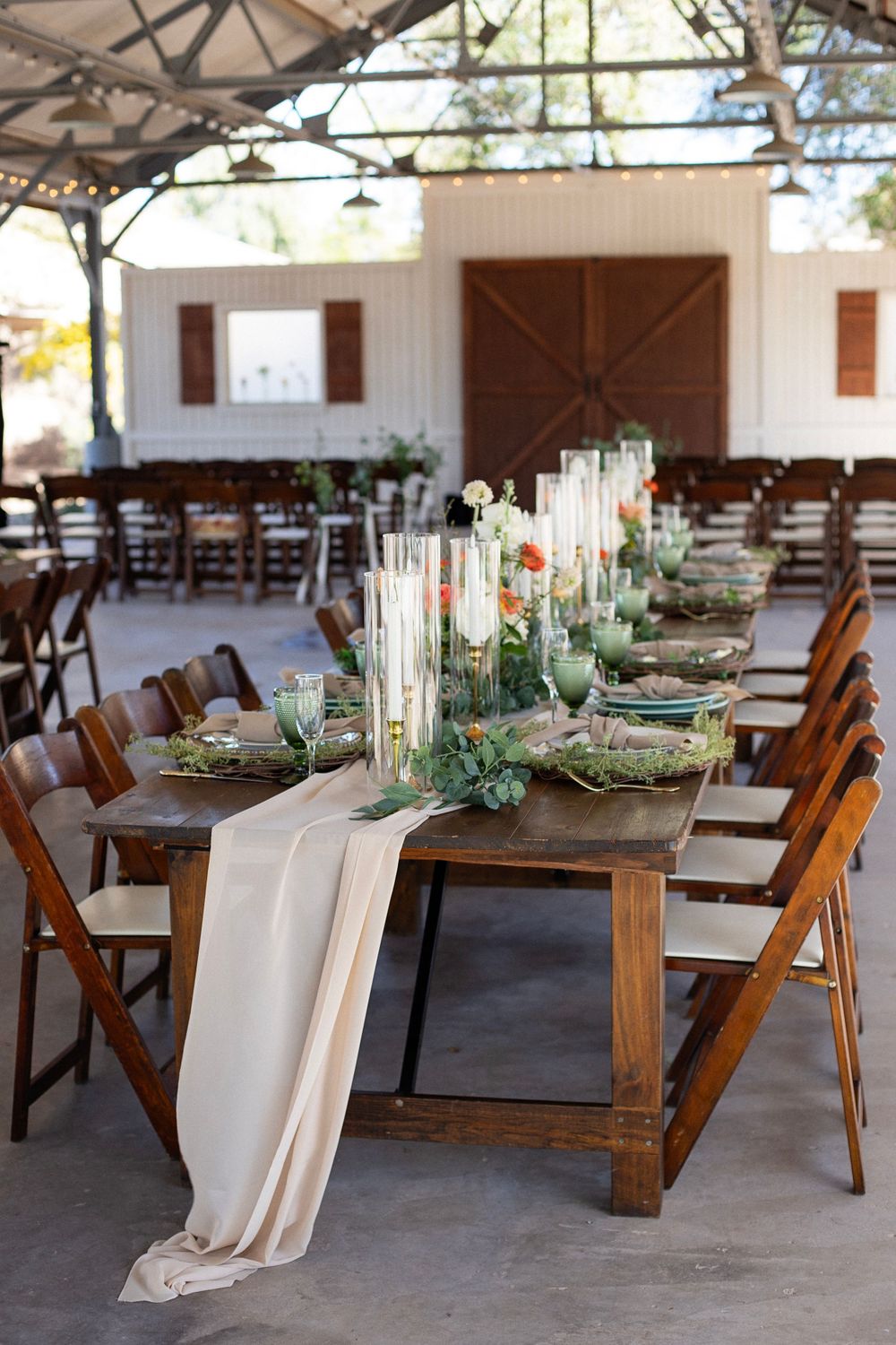 Rustic wedding reception setup with wooden farm tables, greenery runners and white linens.