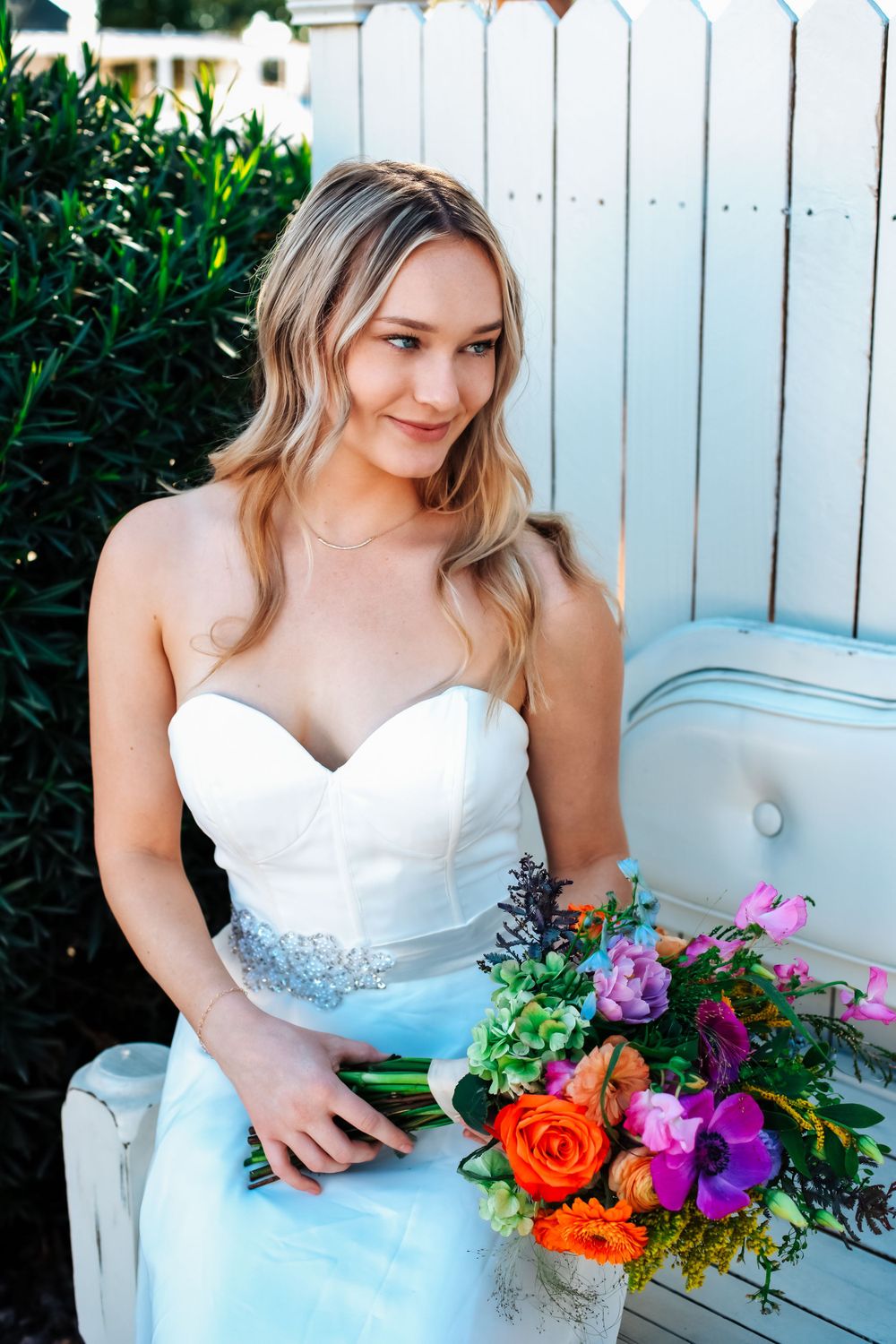 Bride in white dress poses with colorful bouquet on garden bench.