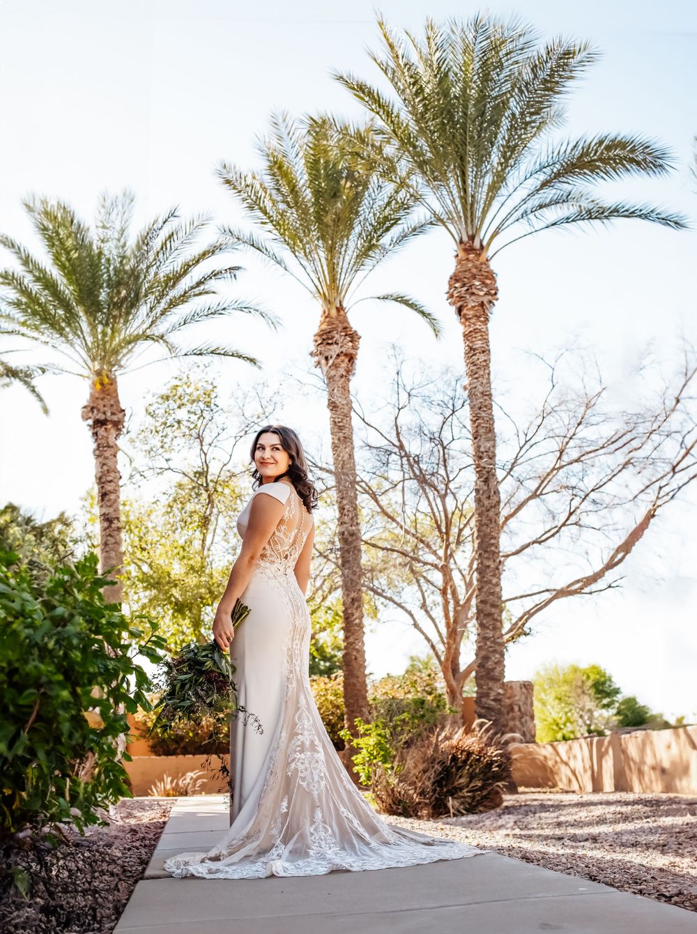 Bride in flowing gown poses elegantly among desert palm trees at sunset.
