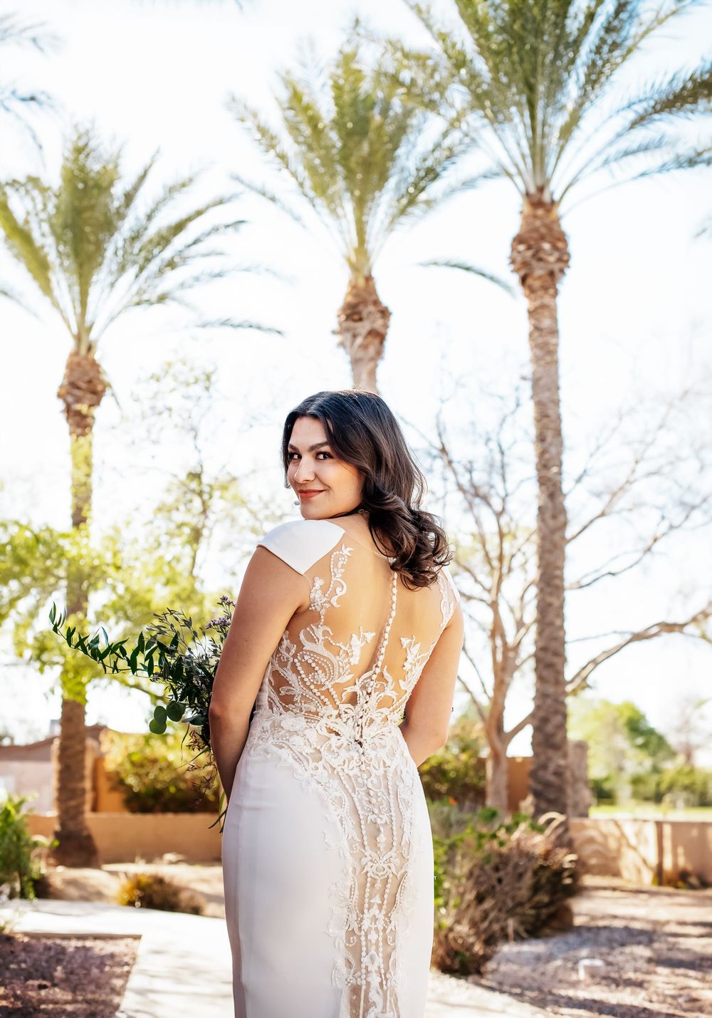 Elegant lace wedding dress details shown against a backdrop of palm trees in a desert garden setting.