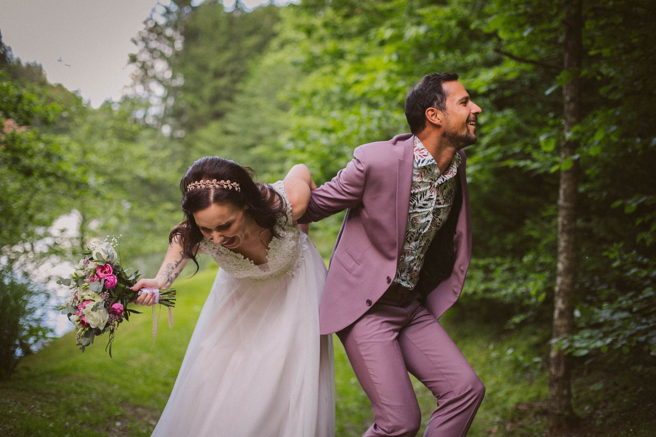 Playful couple in a garden wedding scene with groom in lilac suit and bride holding purple bouquet running together.