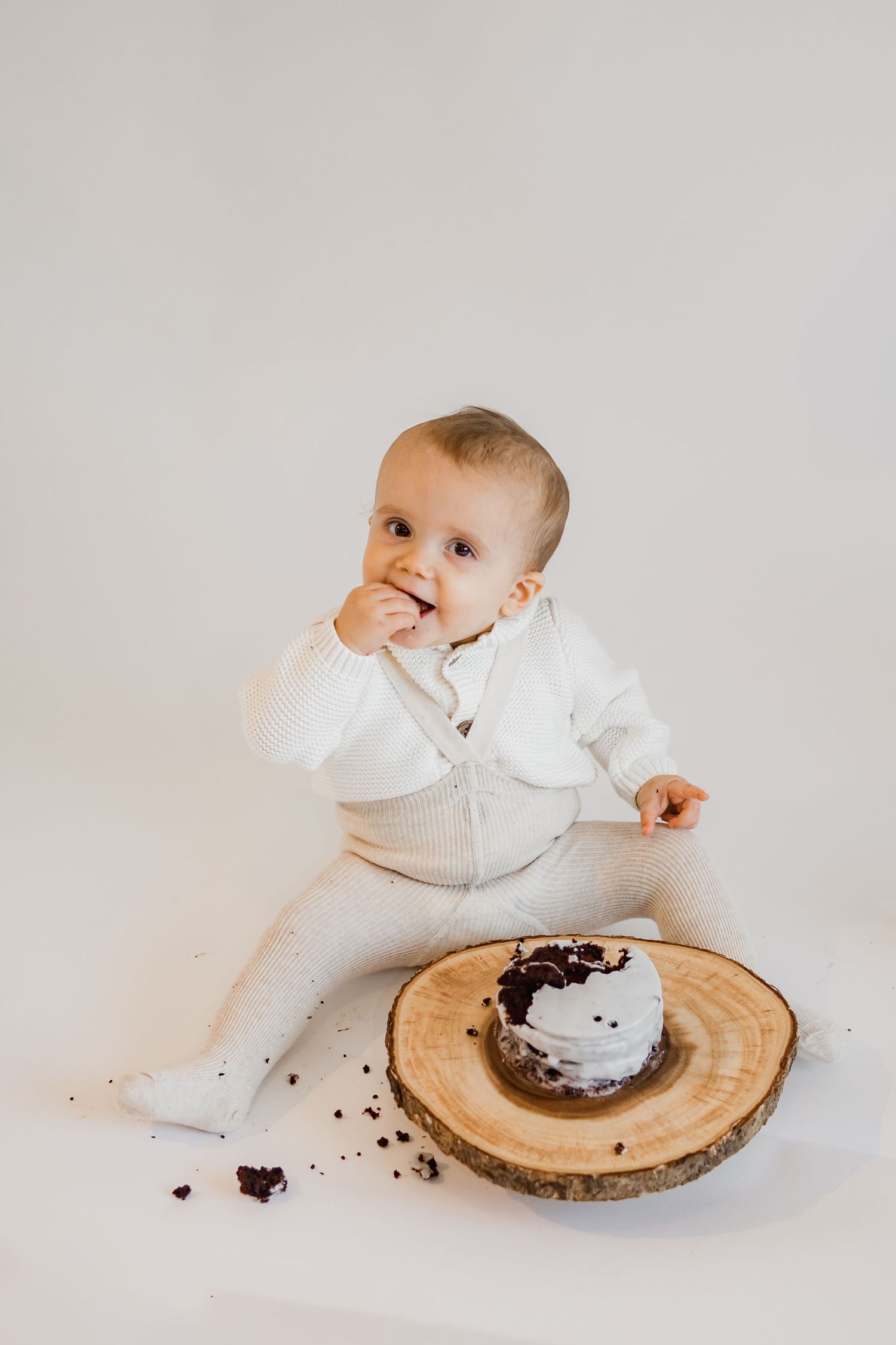 Baby in white outfit sits beside wooden slice with smashed chocolate cake during cake smash photo session.