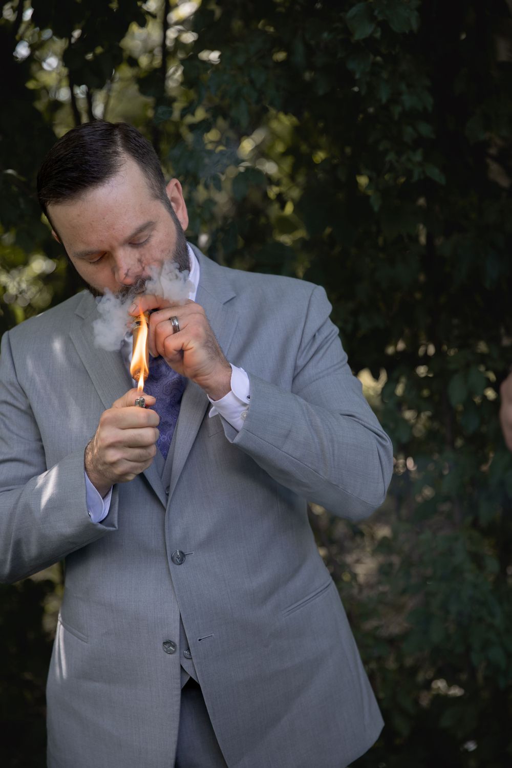 Groom in grey suit lighting a cigar outdoors with smoke billowing.