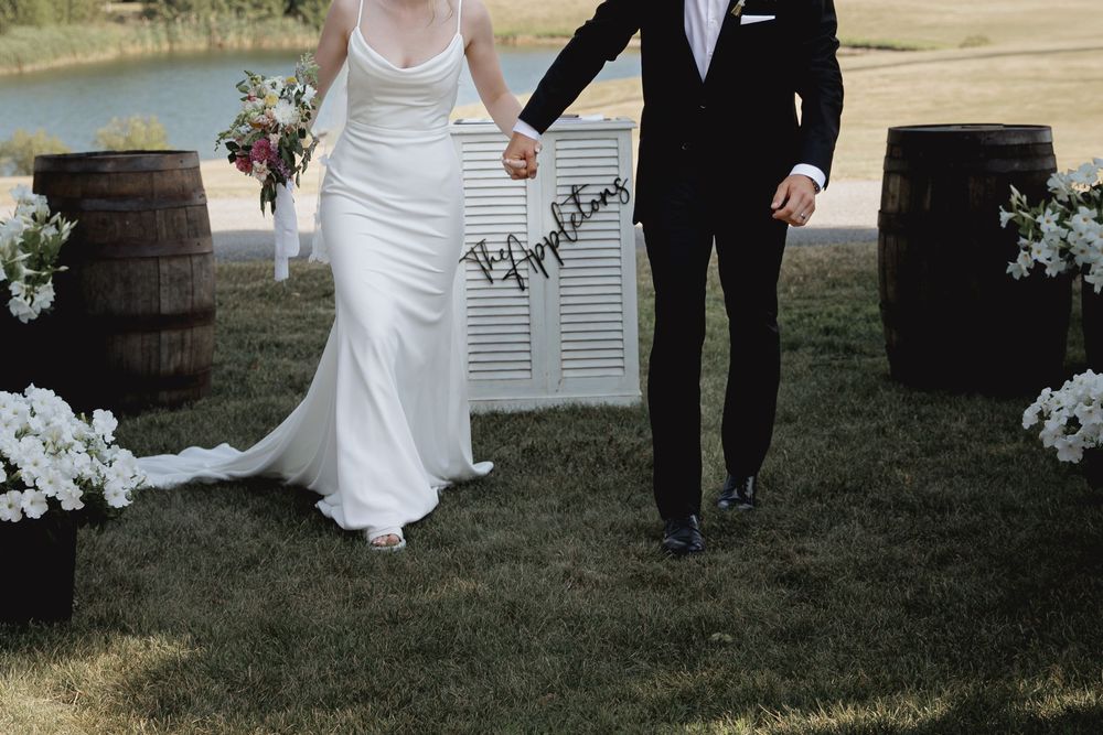 A couple in wedding attire walks together on a grassy outdoor venue with rustic wine barrels in the background.