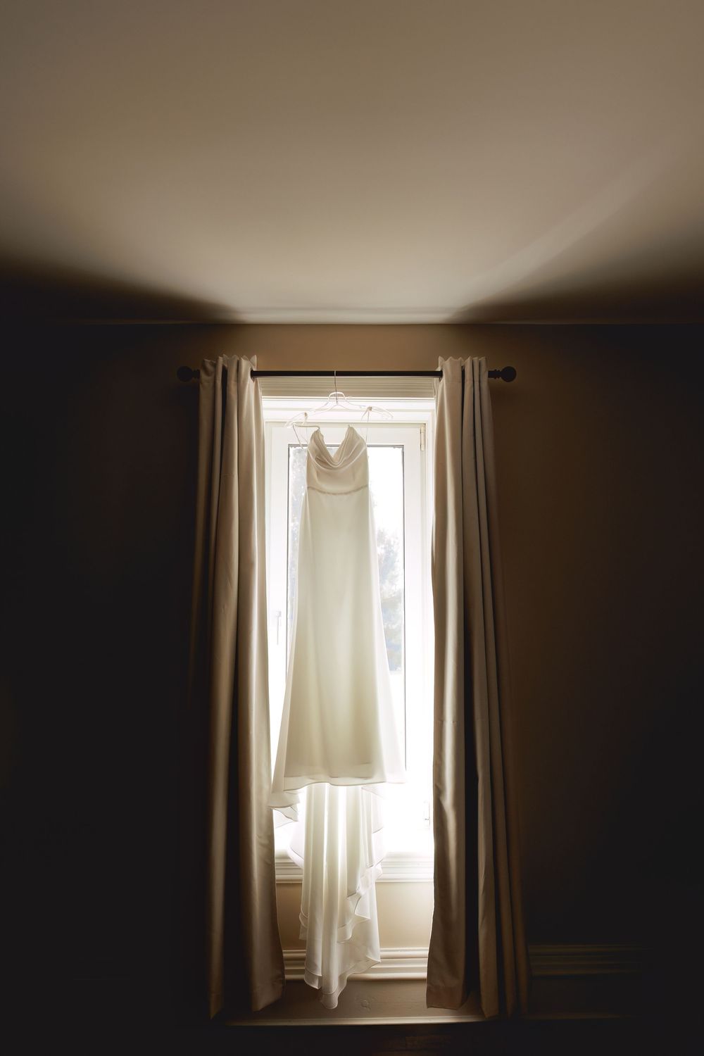 A white wedding dress hangs illuminated in front of a window with dark curtains creating a dramatic silhouette.