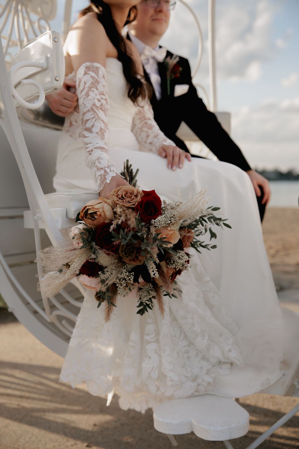 Bride in white lace wedding dress sitting on vintage chair with autumn-toned floral bouquet by the waterfront.