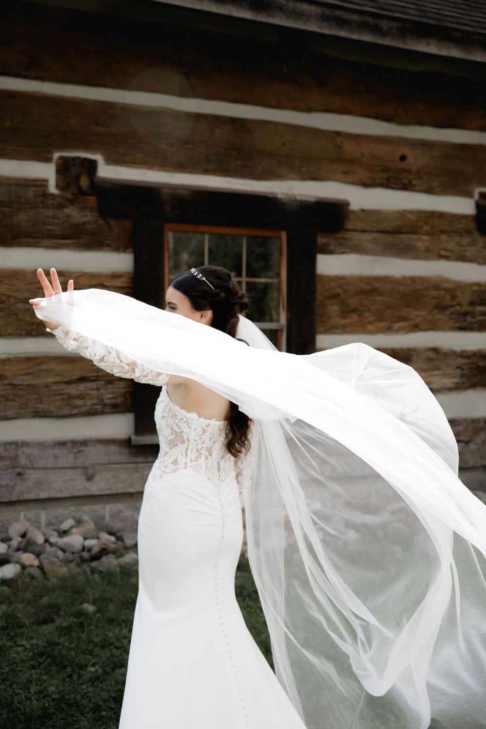 A bride in a flowing white gown and veil twirls gracefully against a rustic log cabin backdrop.