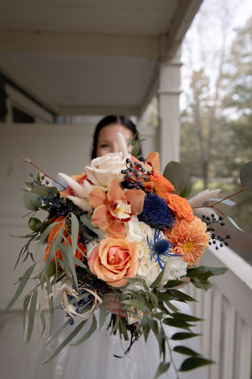 Vibrant bridal bouquet featuring coral roses, blue flowers, and eucalyptus held against white porch railing.