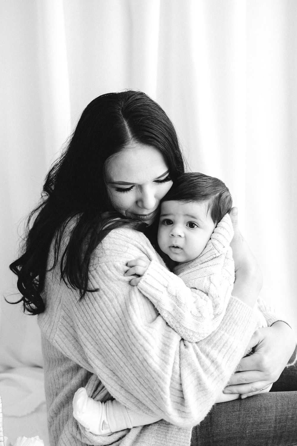 Black and white photo of a loving mother hugging and kissing her baby, both wearing cozy knit sweaters.