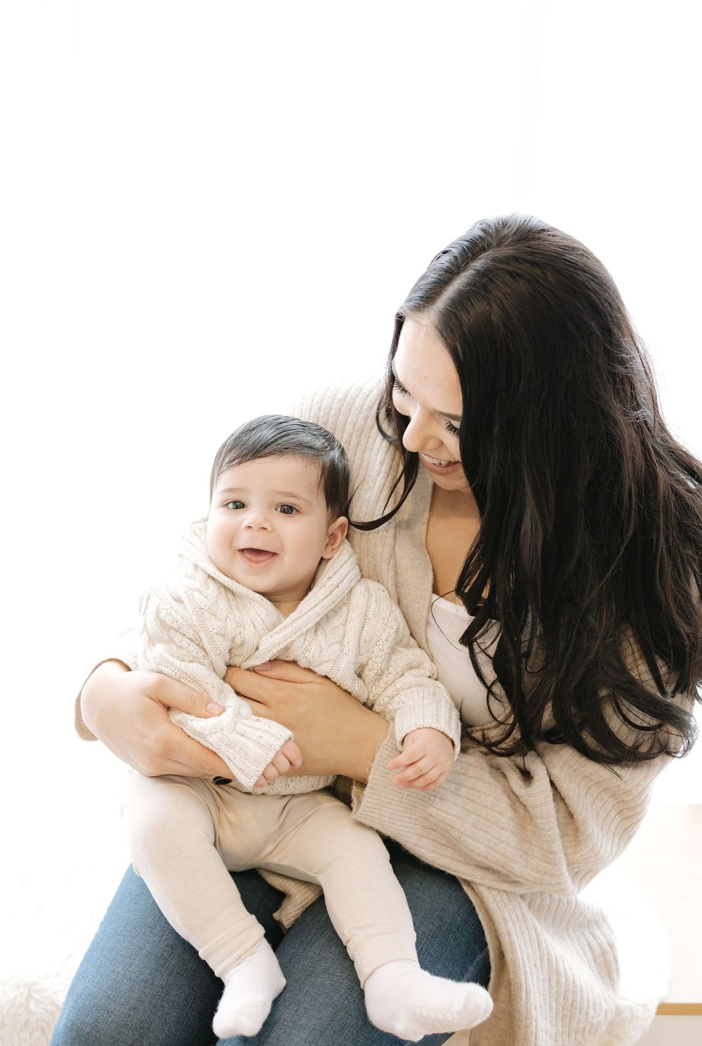 A smiling dark-haired woman holds a happy baby dressed in a cream knit outfit against a bright white background.