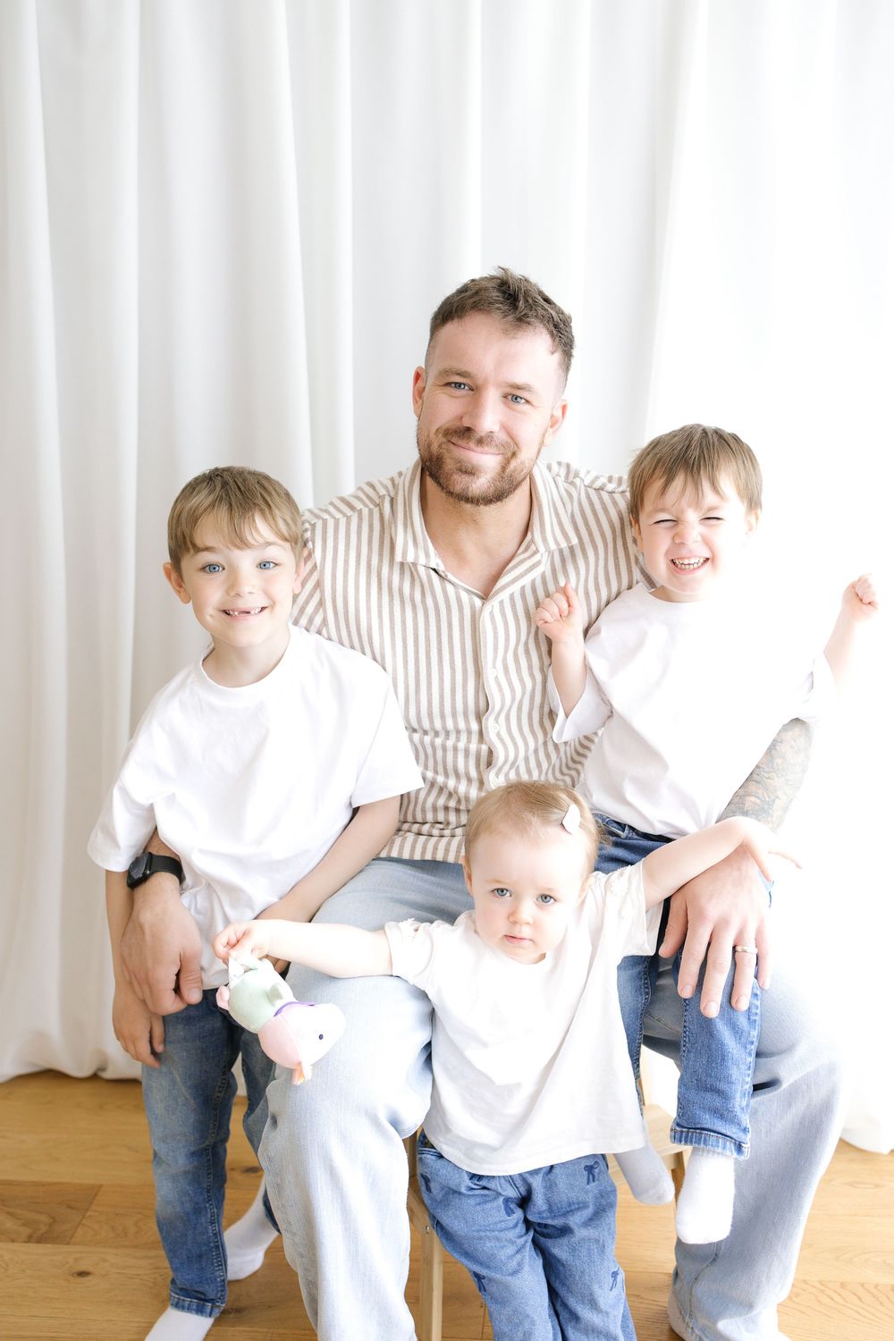Smiling father sitting with three young boys in white t-shirts and jeans posed together for a family portrait.