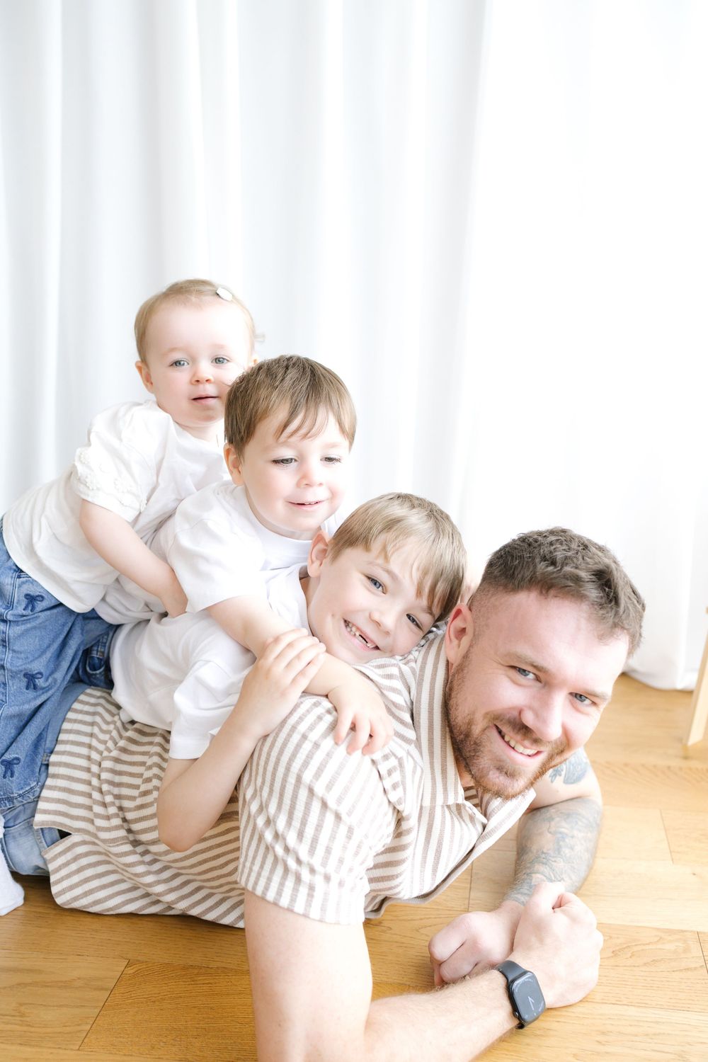 Happy father lying on floor with three young sons stacked on his back, smiling together in bright white room.