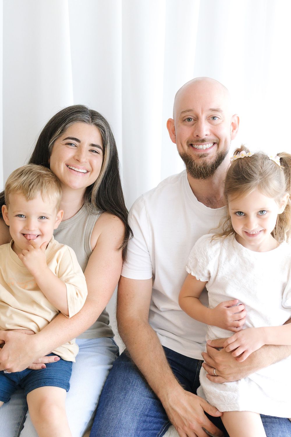 Happy family of four smiling together, parents holding young children in a bright white studio setting.