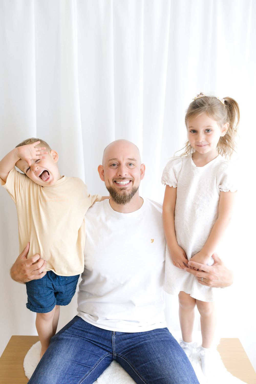 Bald bearded man sitting with two young children, boy and girl, posing together against white curtain backdrop.