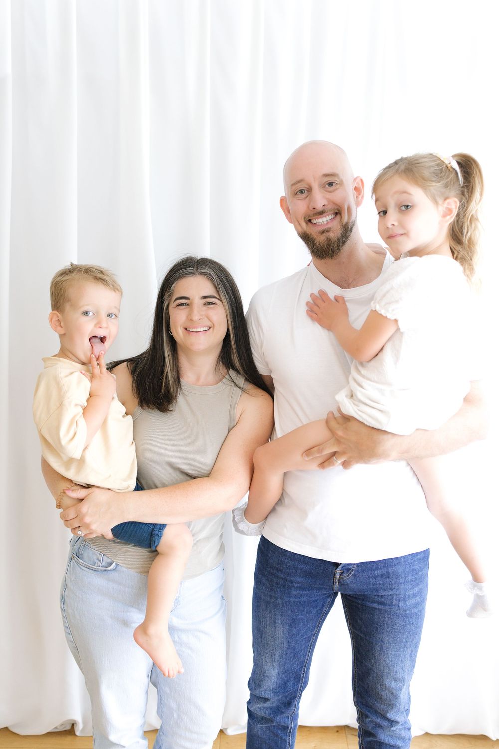 Happy family of four posing together, parents holding two young children, smiling in a bright white studio setting.