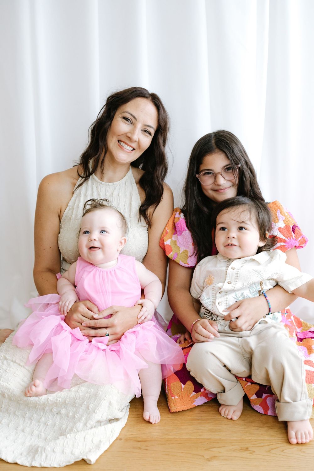 A smiling woman and older girl pose with two babies in pink and white outfits on a wooden floor.