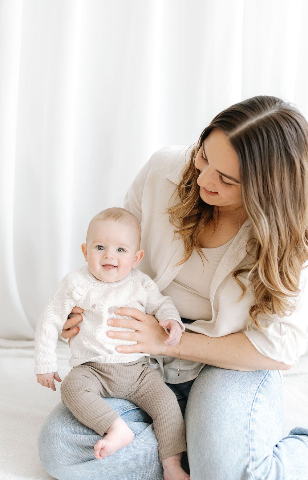 A young mother in a white blazer and jeans holds her smiling baby dressed in a white onesie and tan pants.
