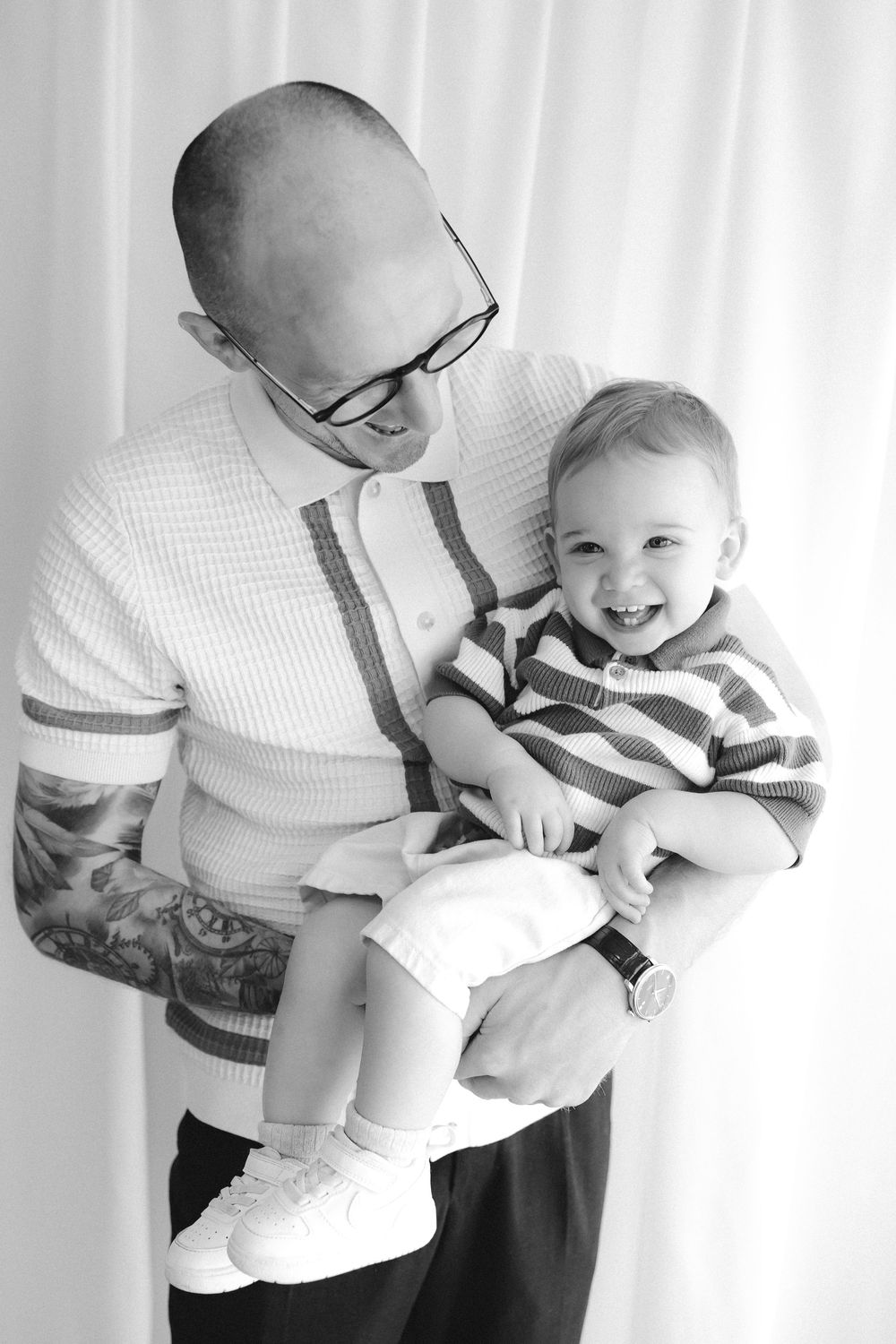 Black and white photo of a bald tattooed man with glasses holding a smiling baby in a striped sweater.
