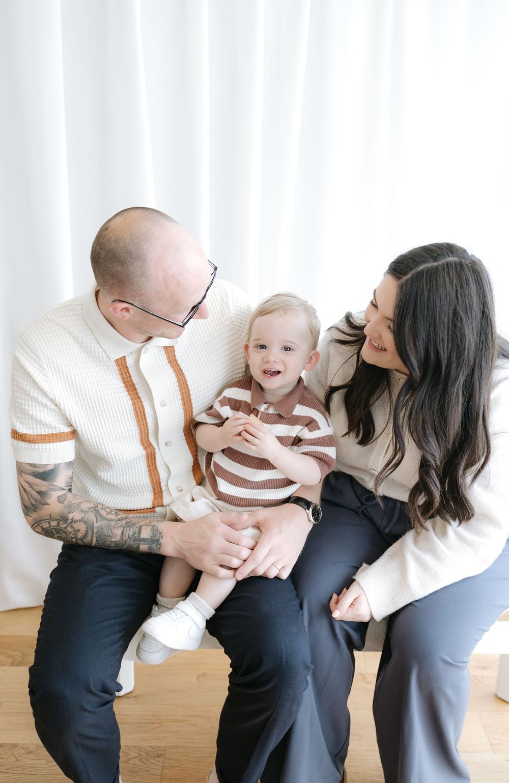 A happy family with tattooed dad, dark-haired mom, and smiling toddler in matching stripes pose in bright white studio.