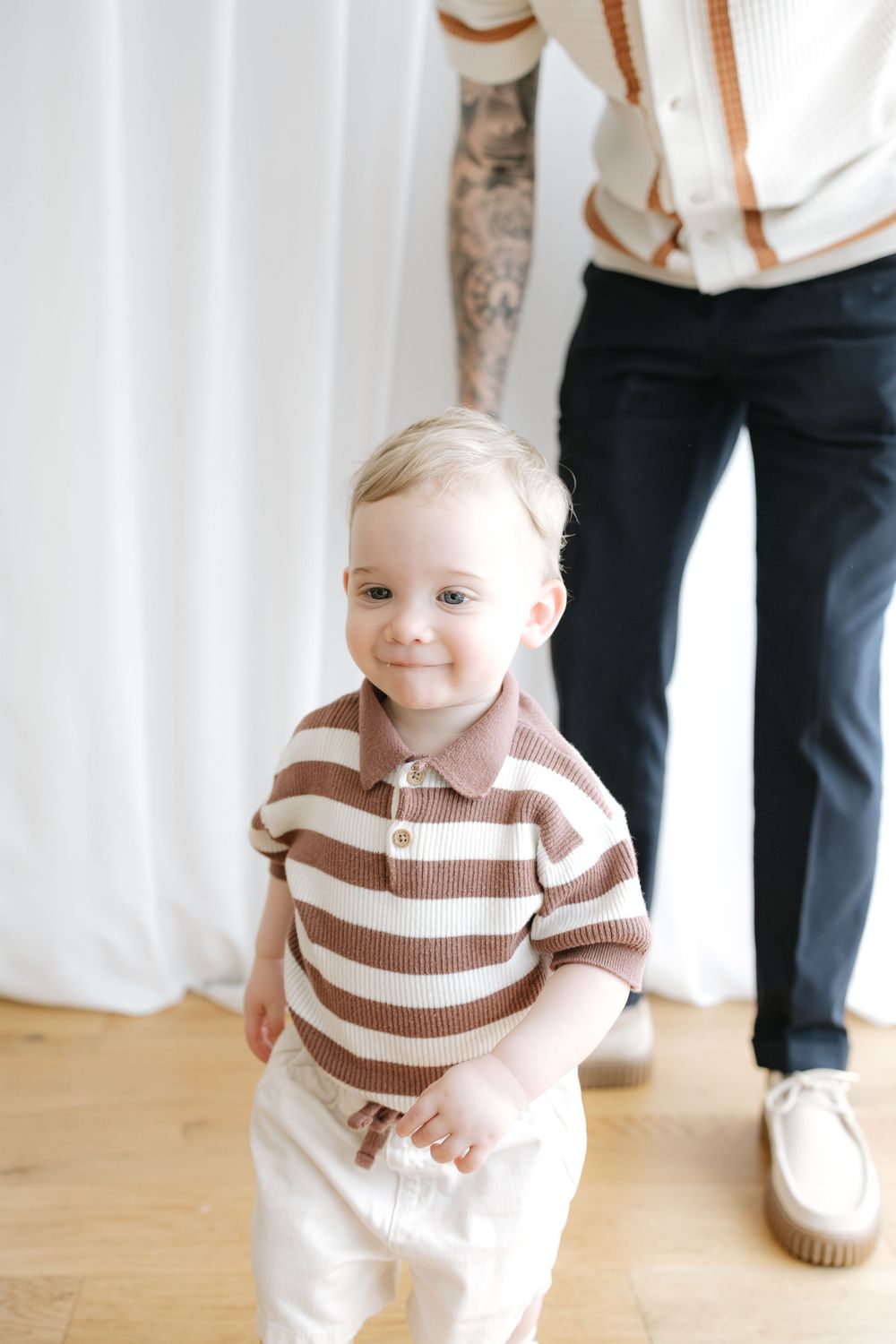 Smiling baby boy in brown striped polo shirt standing on hardwood floor with adult legs visible behind him.