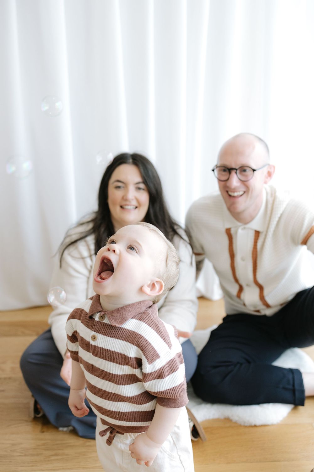 A joyful baby looks up at bubbles while happy parents sit on a wooden floor against a white curtain backdrop.