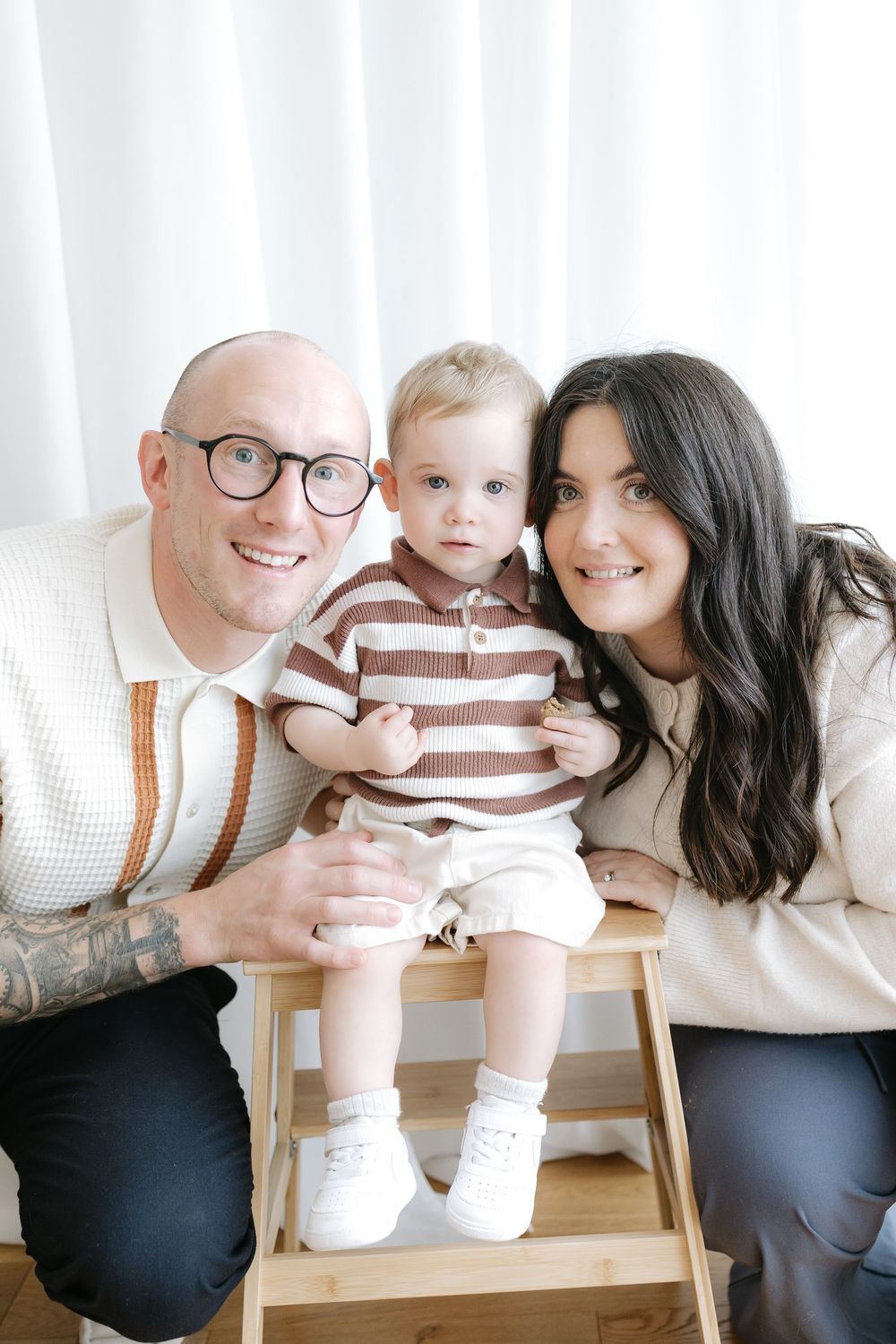A happy family portrait with a bald father in glasses, dark-haired mother, and baby in striped outfit on a wooden stool.