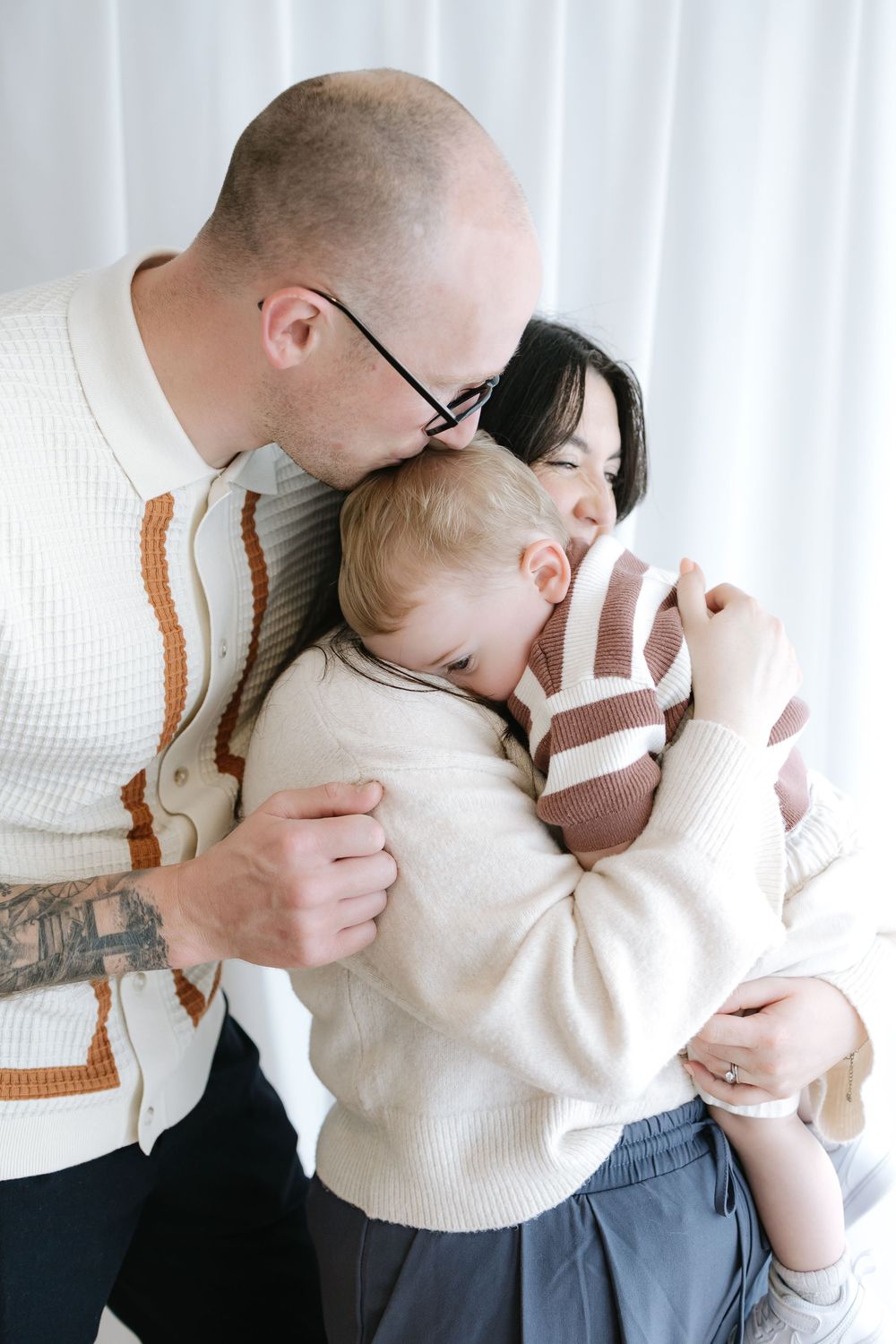 A loving family moment with a bald father kissing his wife as they embrace their toddler in cozy sweaters.