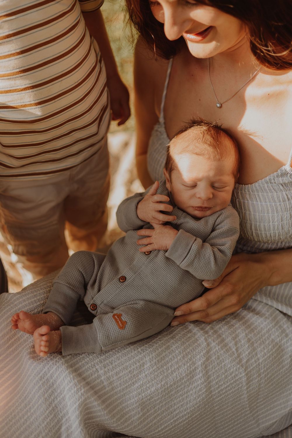 Baby in gray button-up outfit sleeps peacefully in parent's arms in warm-toned intimate portrait.