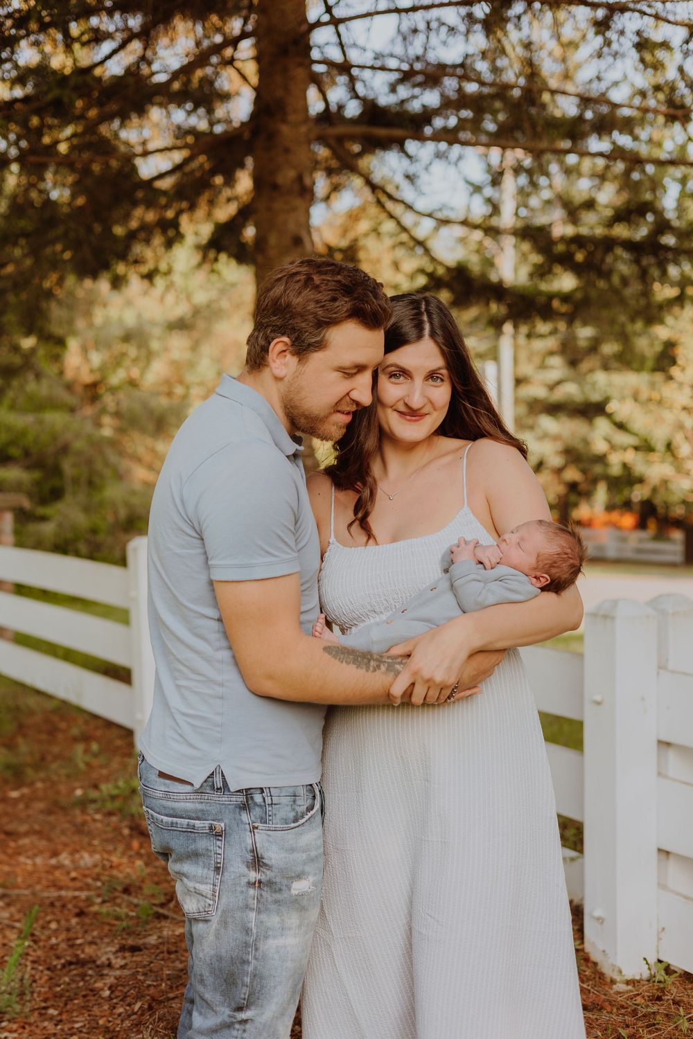 A couple stands outdoors by a white fence at sunset holding their newborn baby in a tender family moment.