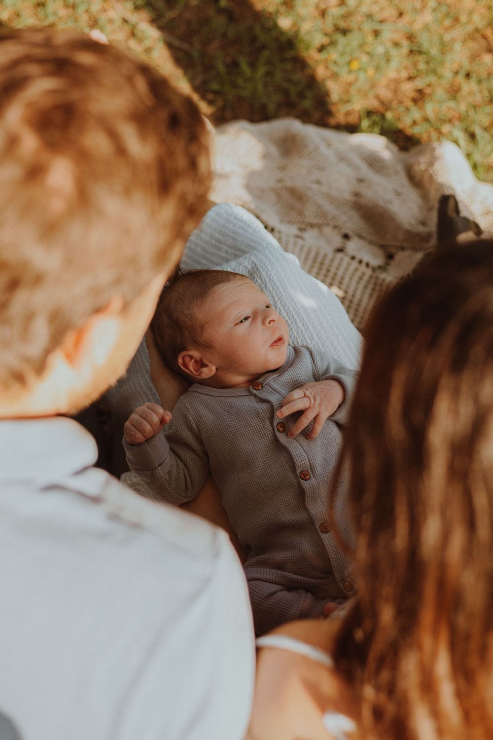 A baby in a gray sweater lays on a blanket while others look on during a warm sunset-lit moment outdoors.