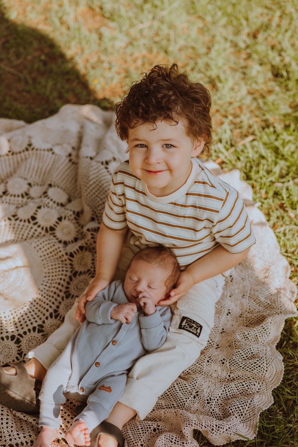 Two young children sitting together on a lace blanket in the grass wearing striped and light colored outfits.