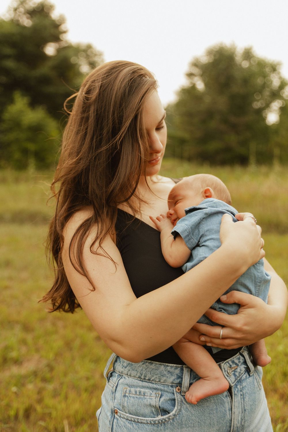 A mother in jeans holds and comforts her baby in a natural outdoor setting during golden hour.