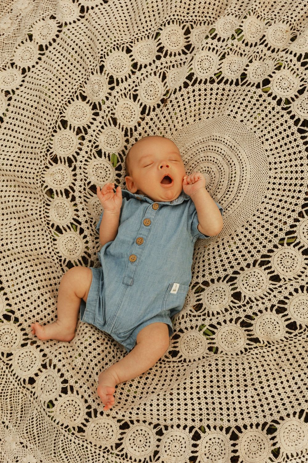 Baby in blue chambray romper yawns while lying on intricate cream crochet blanket with circular lace pattern.