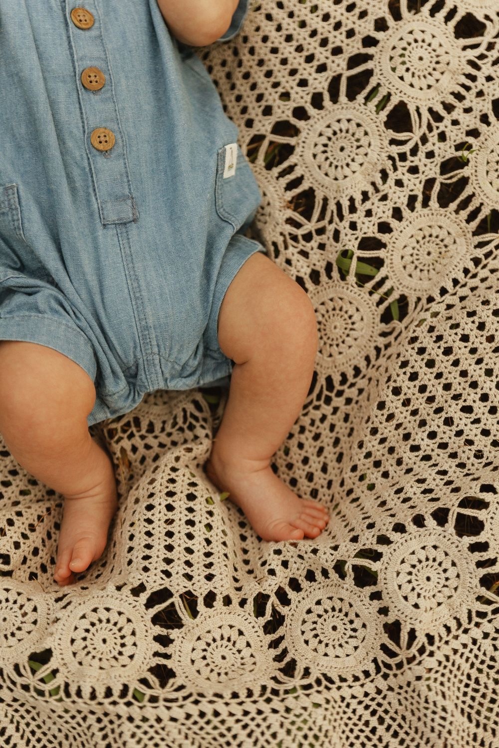 Baby's tiny feet rest on intricate white crocheted lace with a light denim shirt visible in the background.