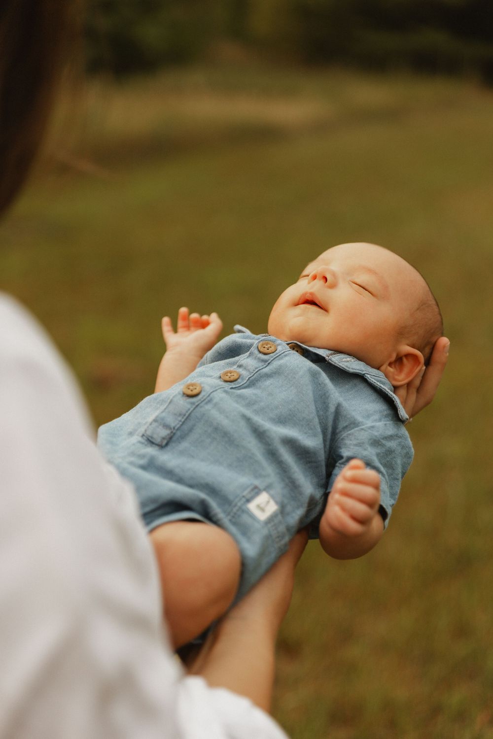 A baby wearing blue overalls rests peacefully outdoors while being held on a sunny day.