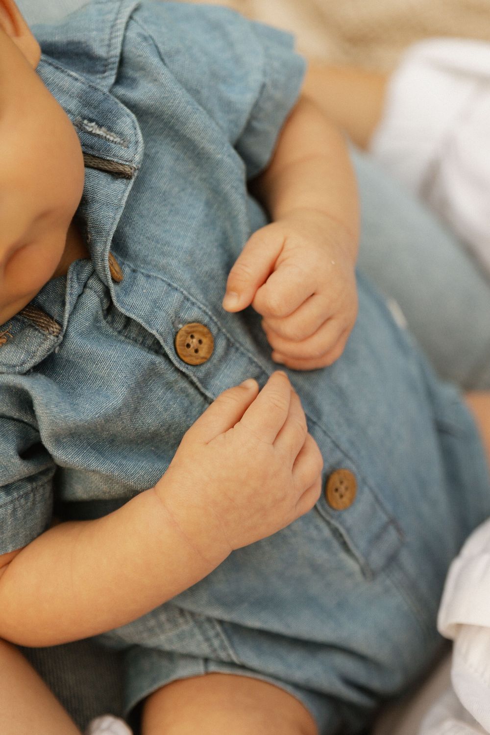 Close-up view of small hands fastening buttons on soft denim clothing against white fabric background.
