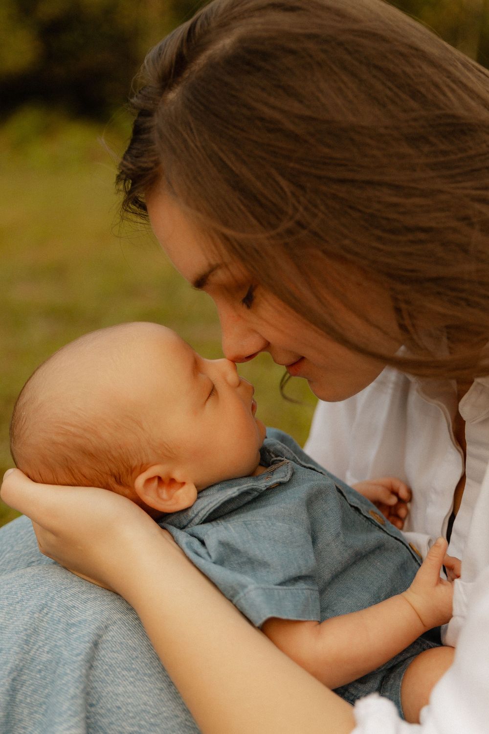 A tender moment between parent and baby in white and chambray, sharing a gentle close embrace outdoors.