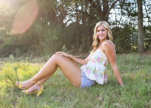 Smiling blonde woman sitting in a sunny green field wearing a floral top and denim shorts.