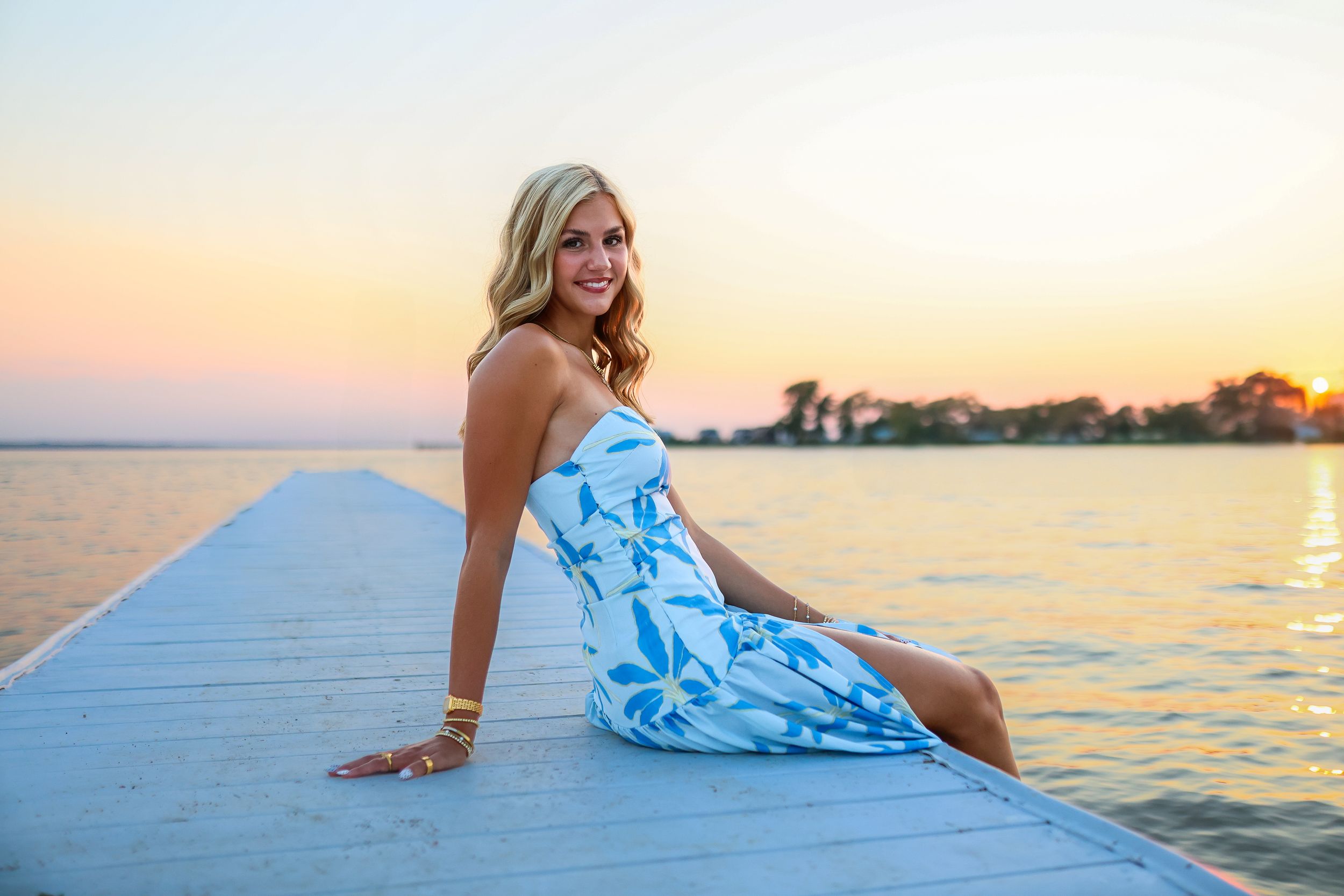 Blonde woman in a blue floral dress sitting on a dock at sunset over calm water.