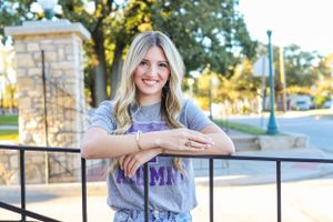 Smiling blonde woman leaning on a railing outdoors wearing a purple alumni graphic tee.