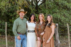 Family of four posing outdoors near a fence with trees, man in cowboy hat and green shirt with three women.