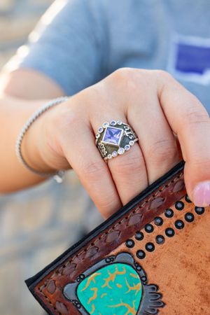 Close-up of hand wearing silver class ring with blue gemstone resting on decorative leather clutch purse.