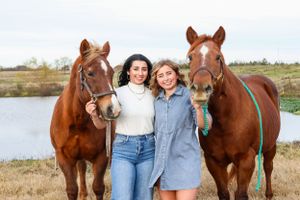 Two young women smile together holding two chestnut horses by a scenic pond outdoors.