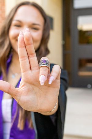 Smiling young woman in purple showing off silver class ring with purple gemstone up close to camera.