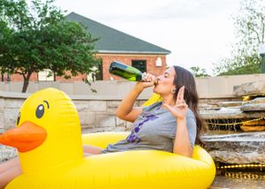 Woman relaxing on giant yellow inflatable duck float drinking wine bottle in outdoor pool setting.
