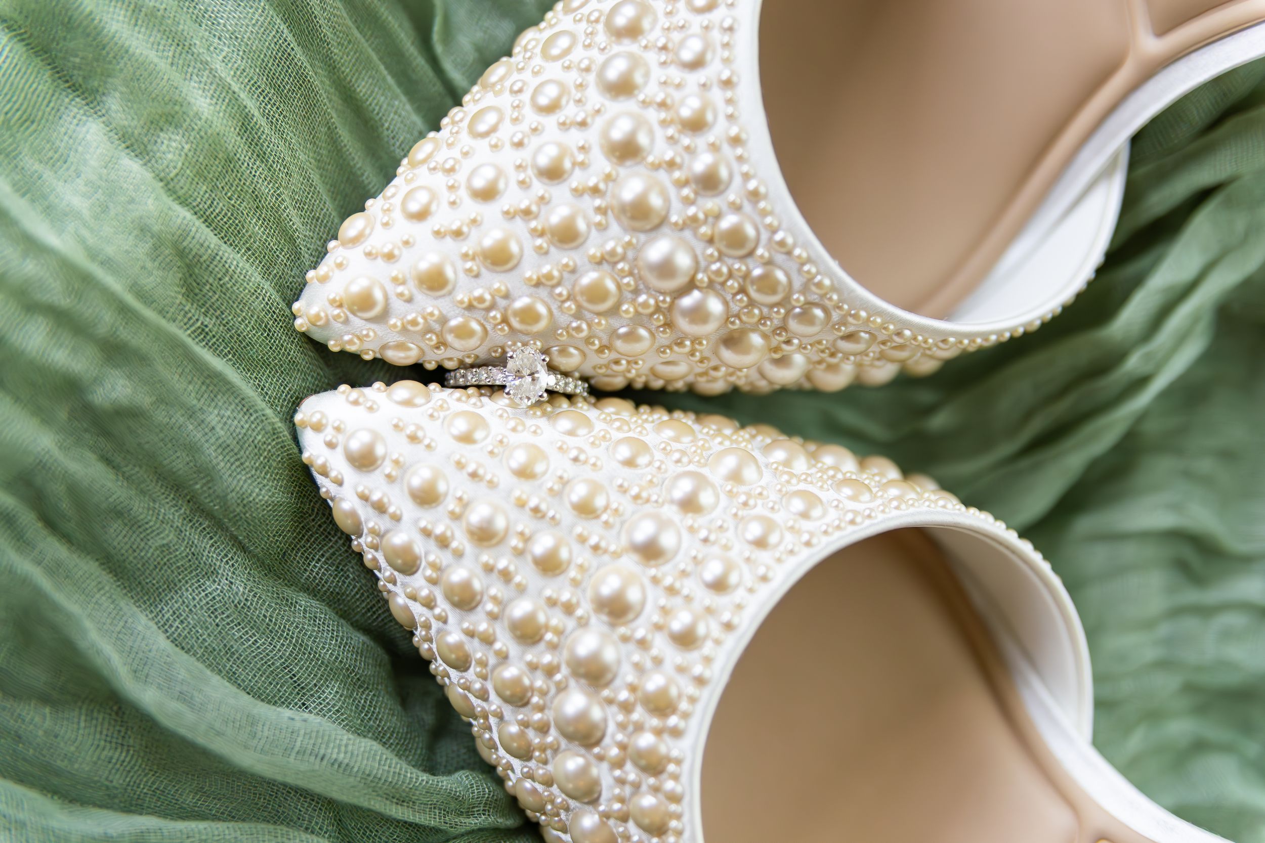 Close-up of elegant pearl-encrusted white wedding heels on green fabric.