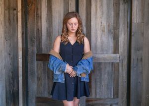 Young woman in navy dress and denim jacket standing before rustic wooden barn doors.